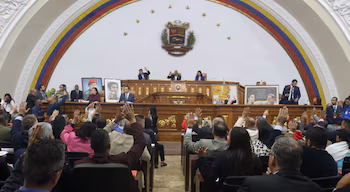 De nombreux législateurs assis lèvent la main lors d'un vote dans une salle de réunion avec une estrade principale et le drapeau vénézuélien