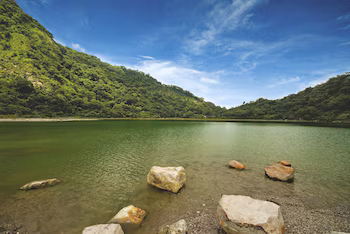 Vue panoramique sur un lac d'eau verte, avec de gros rochers sur le rivage, entouré de montagnes couvertes de végétation sous un ciel bleu nuageux