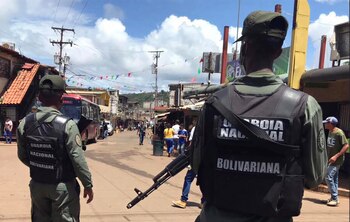 Deux membres de la Garde nationale bolivarienne, dos tournés, portant des uniformes et des gilets militaires, l'un avec un fusil, patrouillent dans une rue remplie de bâtiments et de personnes