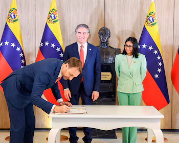 Un homme en costume bleu foncé signe un document à une table blanche, flanqué de deux personnes et de drapeaux vénézuéliens dans un salon.