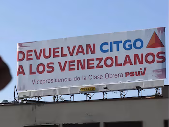 Un panneau d'affichage blanc avec le message "RETOURNER CITGO AUX VÉNÉZUELENS" en lettres rouges et bleues, et le logo du PSUV, sous un ciel bleu clair