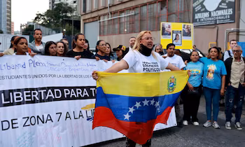 Une femme avec des lunettes et un t-shirt blanc qui dit "Liberté pour tous les prisonniers politiques" tient un drapeau du Venezuela devant un groupe de manifestants avec des banderoles