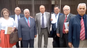 Un groupe de sept avocats, six hommes et une femme, debout devant un immeuble. La plupart d’entre eux portent des vêtements formels et certains tiennent des documents à la main.