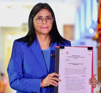 Une femme aux cheveux noirs et portant des lunettes, vêtue d'un blazer bleu, tient un document avec un texte rouge et noir indiquant « LOI D'AMNESTÉ ».