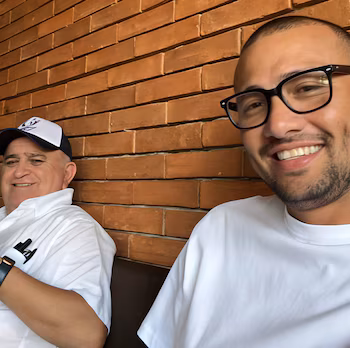 Deux hommes souriants assis ; un plus âgé avec une casquette et une chemise blanche, et un jeune avec des lunettes et un t-shirt blanc. fond de mur de briques