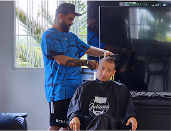 Un coiffeur avec une casquette et une chemise bleue coupe les cheveux de Cheremo, qui porte une cape noire avec le logo 