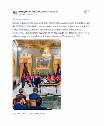 Un homme en costume sombre signe des documents à une table avec des drapeaux du Venezuela. D'autres participants regardent dans une salle formelle avec un lustre et une peinture représentant un cavalier.