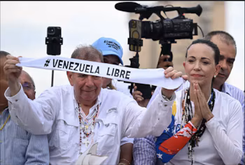 Un homme et une femme tiennent une bannière blanche avec le texte « #VENEZUELA LIBRE ». En arrière-plan, un caméraman avec une caméra de télévision et un téléphone portable