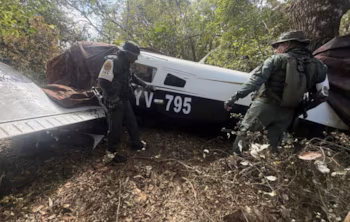 Deux hommes en uniforme inspectent un petit avion noir et blanc immatriculé YV-795, partiellement recouvert d'une bâche, dans une forêt dense
