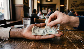 Gros plan d'une main donnant deux billets d'un dollar à une autre main tendue, sur une table en bois dans un restaurant avec des gens en arrière-plan