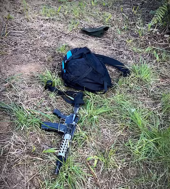 Un fusil d'assaut avec un chargeur, un sac à dos noir avec des détails bleus et une casquette vert foncé posé sur le sol recouvert d'herbe et de terre sèche