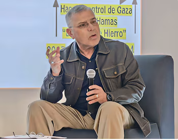 Un homme aux cheveux gris et portant des lunettes, vêtu d'une veste sombre et d'un pantalon kaki, est assis et parle dans un microphone ; derrière, un tableau noir avec un texte sur Gaza