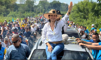 Femme souriante en chapeau de paille et chemise blanche assise sur le toit d'une voiture, saluant une foule sur une route avec des arbres verts en arrière-plan