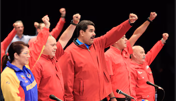 Nicolás Maduro, en veste rouge, et d'autres officiels en chemise rouge lèvent le poing droit sur une scène sombre, avec des microphones au premier plan