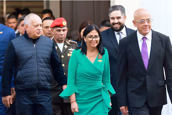 Jorge Rodríguez avec un gilet sombre et Delcy Rodríguez avec une robe verte et des lunettes, marchent en souriant aux côtés d'autres hommes en costume et d'un soldat en uniforme