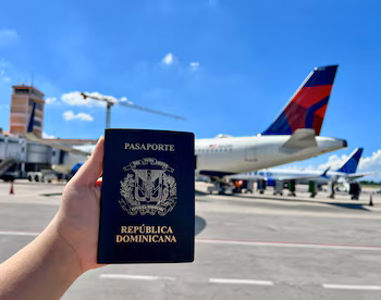 Une main tient un passeport dominicain noir avec un bouclier doré devant la queue rouge et bleue d'un avion Delta sur le tarmac de l'aéroport international du Cibao