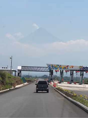Fourgonnette sombre sur route pavée vers une place de péage moderne aux motifs géométriques. Un grand volcan nébuleux s'élève au-dessus des nuages au loin
