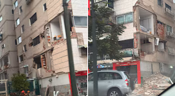 Immeuble à plusieurs étages avec façade endommagée, balcons cassés, briques apparentes, tas de gravats au sol, arbre, poteau et voiture argentée