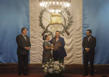Quatre personnes sur scène avec le drapeau du Guatemala et ses armoiries. Deux femmes au centre, l’une tend un cadre à l’autre. Composition florale et candélabre