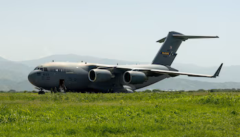Grand avion militaire gris avec inscription US AIR FORCE, quatre moteurs, porte d'entrée ouverte, personnel militaire, herbe et montagnes lointaines