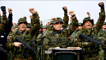 Groupe de soldats avec uniforme de camouflage et armes, avec plusieurs poings levés. Un officier devant parle sur un podium avec un microphone. Quelques patchs avec texte "FANB"