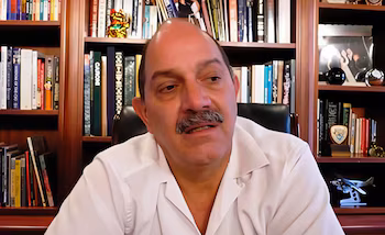 Homme à la peau claire avec moustache, vêtu d'une chemise blanche, assis devant une étagère en bois avec des livres et des ornements