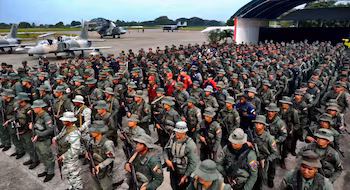 Un grand groupe de soldats en uniforme, camouflés et fusils, se forme sur une piste d'atterrissage avec des avions de combat et un hélicoptère en arrière-plan.