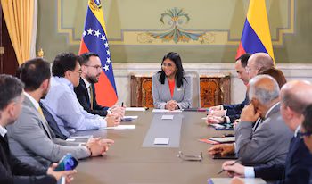 Une femme souriante vêtue d'un blazer gris et d'un chemisier rouge est assise à une table de conférence avec plusieurs hommes. Drapeaux du Venezuela et de la Colombie en arrière-plan