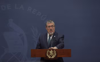 Un homme en costume-cravate prononce un discours depuis un podium en bois. Derrière, un mur bleu avec les armoiries du Guatemala et le texte 'DE LA REPÚ'