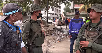 Deux soldats avec des uniformes et des chapeaux verts, un civil avec une casquette et un masque, un autre civil avec une chemise bleue. Débris, pelle rétrocaveuse et personnes en arrière-plan