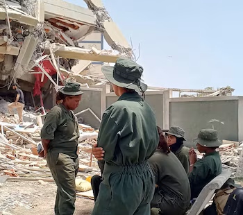 Plusieurs femmes en uniforme militaire vert et chapeaux verts sont debout ou assises à côté d'un bâtiment effondré et de décombres sous un ciel dégagé