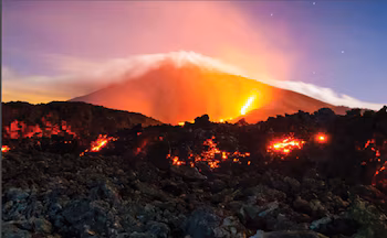 Vue nocturne du volcan Fuego en éruption avec de la lave incandescente coulant sur ses pentes et un panache de cendres au sommet, sous un ciel étoilé