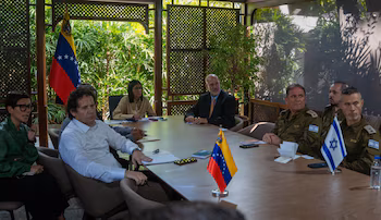 Groupe de personnes assises autour d’une table en bois. Des drapeaux du Venezuela et d'Israël sont visibles, ainsi que des hommes en uniforme militaire et des personnes en civil.