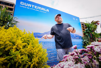 Homme barbu portant une casquette, un t-shirt et un short devant une bannière bleue qui dit « GUATEMALA LAKE ATITLÁN, SOLOLÁ ». Arbustes et fleurs au premier plan
