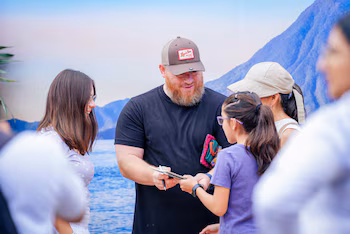 Un homme barbu coiffé d'une casquette signe un cahier à une fille à lunettes. Une autre femme sourit à gauche. Fond de paysage de montagne et d'eau peint