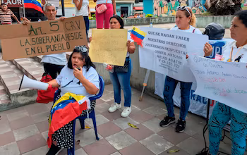 Plusieurs personnes sur une place brandissent des pancartes et des drapeaux du Venezuela, une femme assise parle dans un mégaphone. Les panneaux contiennent du texte en espagnol