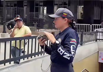 Femme portant une casquette grise et un uniforme bleu foncé, avec les emblèmes et le drapeau du Venezuela, tient un objet gris. Un homme avec un polo jaune en arrière-plan