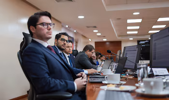 Des hommes en costume assis à une table. Il y a des ordinateurs portables, des moniteurs, des microphones, des tasses et des lunettes. Un panneau visible indique « Javier Andres Alfaro Fitch Ratings »