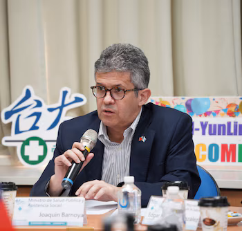 Homme avec des lunettes, un costume et un épinglette du drapeau guatémaltèque, parlant dans un microphone. Sur la table, une assiette avec « Joaquin Barnoya », des bouteilles d'eau, des verres à café. Fond avec texte chinois
