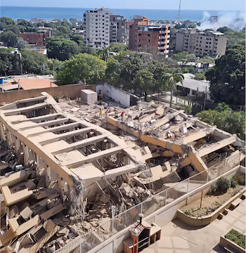 Bâtiment partiellement effondré avec des décombres, trois personnes dessus, d'autres bâtiments et arbres en arrière-plan, mer et ciel dégagé à l'horizon