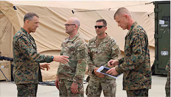 Quatre hommes en uniforme militaire, deux en camouflage vert foncé et deux en camouflage vert clair, discutent devant des tentes