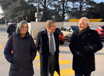 Trois adultes, deux hommes et une femme, debout à l’extérieur, avec des arbres et des véhicules en arrière-plan. Le sol a des lignes jaunes