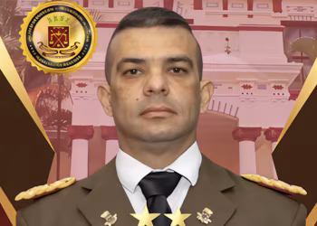 Portrait en buste d'homme en uniforme militaire marron, chemise blanche, cravate noire. Badges dorés sur les revers et les épaules. Emblème doré. Contexte architectural