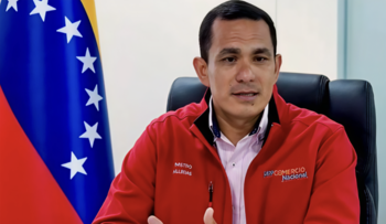 Homme assis, porte une veste rouge avec des badges "M. Allcrias" et "DPP Commerce National" sur la chemise. A votre gauche, le drapeau du Venezuela