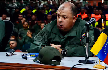 Officier militaire en uniforme vert olive avec des écussons FANB et REDI Los Llanos assis à une table avec microphone, drapeau du Venezuela, casquette et lunettes