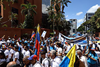 Une foule de gens défilent dans une rue urbaine avec des immeubles et des palmiers. Ils portent des drapeaux vénézuéliens et une banderole à l'effigie de María Corina Machado