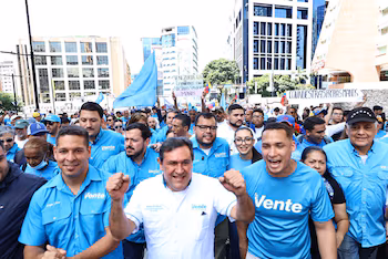 Une foule de gens marchent le long d’une rue urbaine avec de grands immeubles. Plusieurs manifestants portent des t-shirts bleu clair avec "Viens" et ils tiennent des drapeaux
