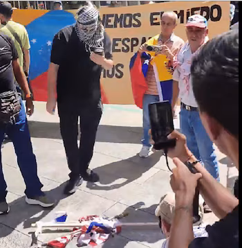 Groupe de personnes dans un espace extérieur, certaines tenant un drapeau du Venezuela, une bannière jaune avec du texte, deux caméras et des morceaux de tissu de drapeau au sol