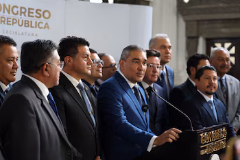 Groupe d'hommes en costumes formels, l'un d'eux s'exprimant depuis un podium avec un microphone. En arrière-plan, un mur avec le texte Congrès de la République