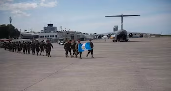 Des soldats en uniforme marchent sur une piste d’aéroport. Certains portent le drapeau du Guatemala. Un grand avion cargo et des bâtiments aéroportuaires visibles